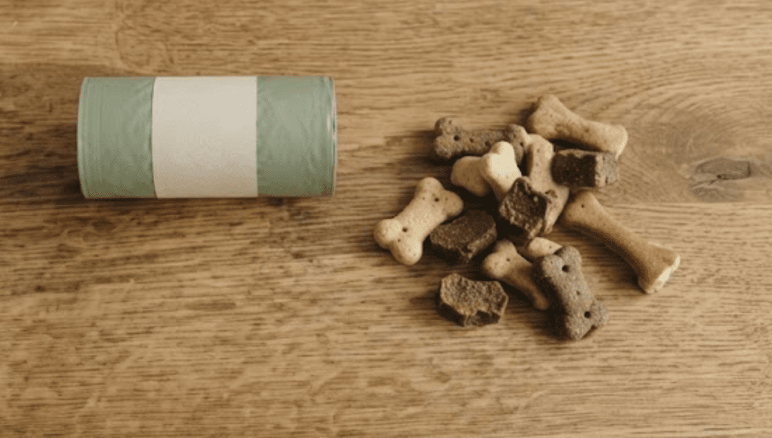 A refill tin of poop bags beside a scattering of dog biscuits on a wooden table.